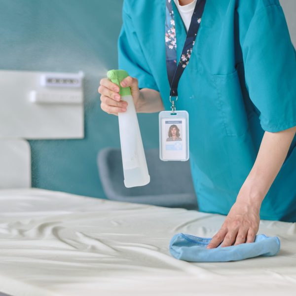 Middle aged Caucasian woman cleaning hospital bed with spray bottle and cloth, wearing medical uniform and identification badge, performing sanitation in healthcare facility